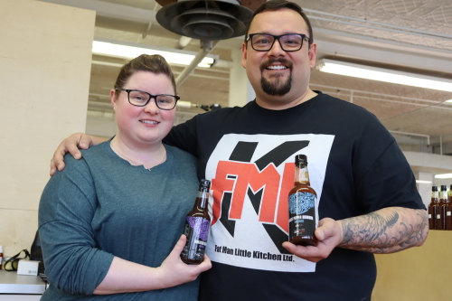 Owners of Fat Man Little Kitchen Samantha Boomer and Marcel Martell stand together at the farmers market holding hot sauce bottles.