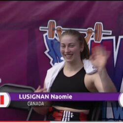 Naomie Lusignan waves at the camera wearing a weightlifting singlet in front of a purple background.