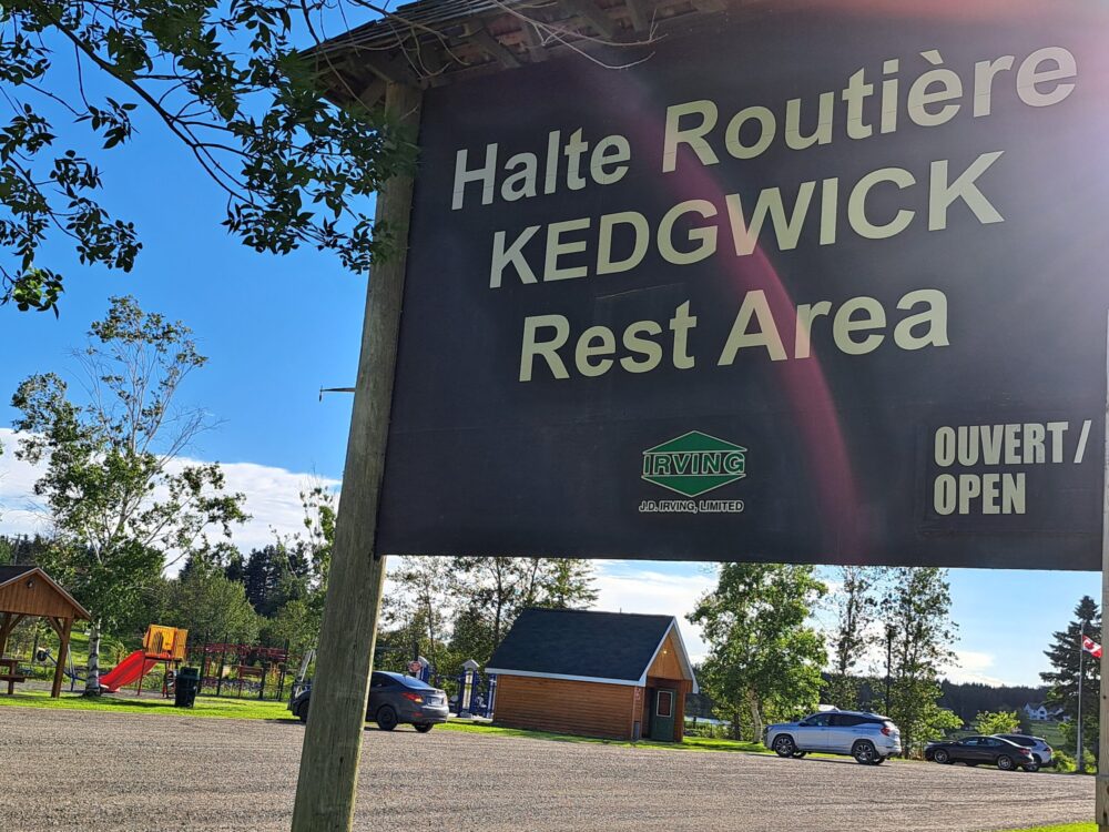 Pancarte affichant la halte routière de Kedgwick avec trois automobiles stationnées devant le parc et par une belle journée dégagée