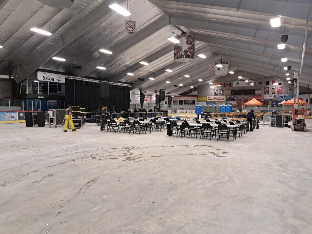 Sur une vaste plancher de ciment à l'intérieur d'un aréna, se trouve des tables et une grande scène de spectacle. Quelques travailleurs procèdent à des installations