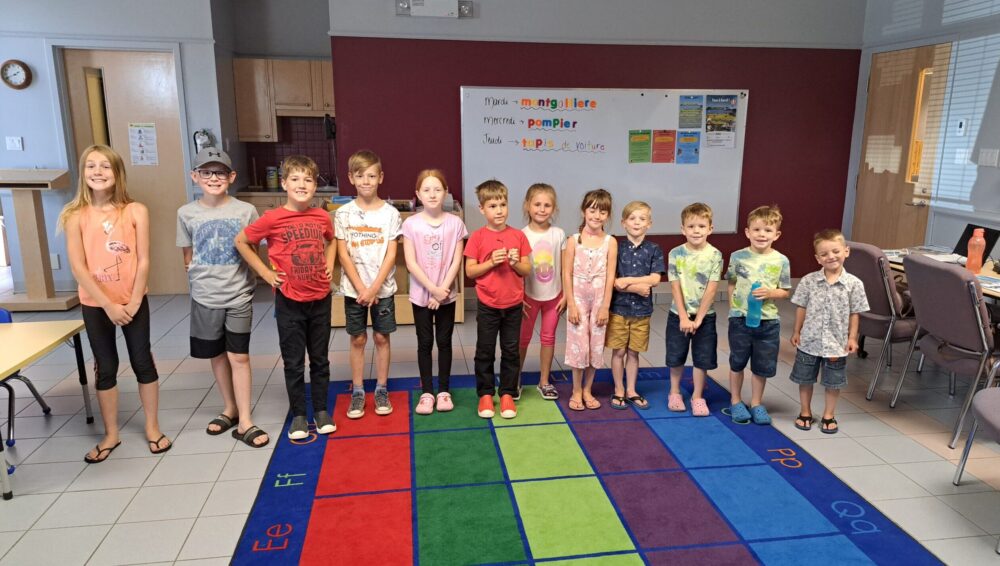 Une dizaine d'enfant en ordre de grandeur de gauche à droite, sur un tapis de carrés de couleurs diverses, dans la pièce d'une bibliothèque