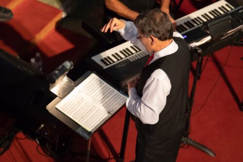 Un homme dirige une chorale en arrière d'un lutrin et d'un piano.