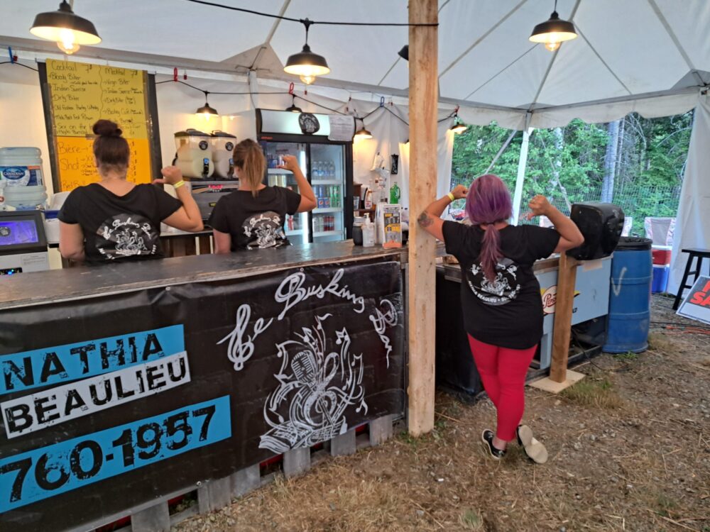 Trois femmes à dos tourné pointant vers le logo de leur chandail noir, derrière le bar d'un festival.