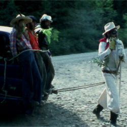 A man in a white hat and suit appears to be hauling a vehicle on foot with three people riding on top.