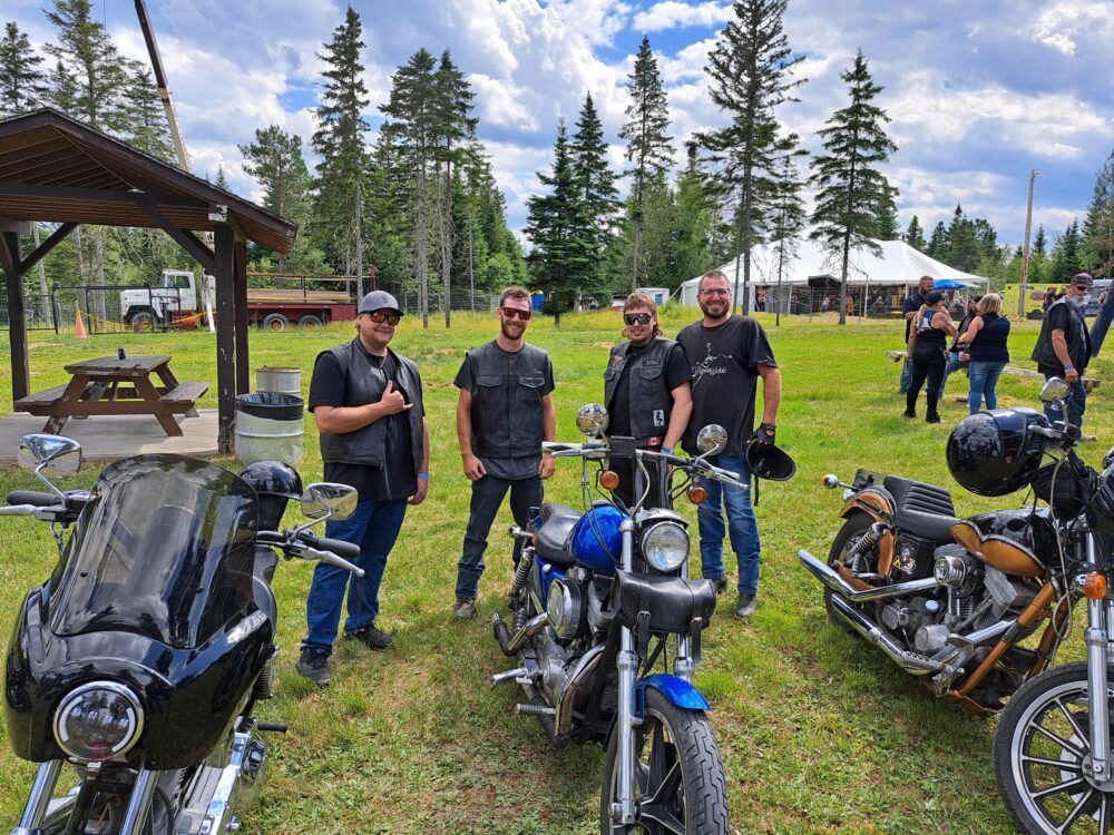 Quatre hommes habillés en motard, tout sourire derrière leur moto sur du gazon, lors d'une journée ensoleillée.