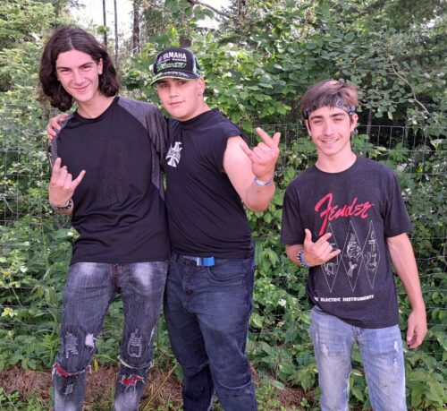Trois adolescents tous vêtus de noir regarde la caméra en souriant devant un boisé lors d'une journée ensoleillée