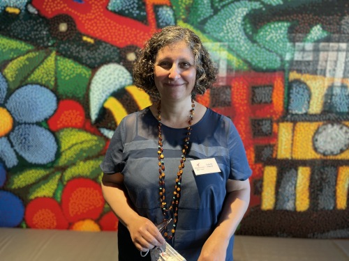 Isabelle Pedot stands in front of a colourful mural on a wall. She is wearing a blue t-shirt and holds a mask in her hand.