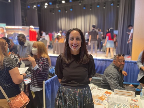 A woman smiling at a job fair. Behind here, there are booths and there is a large stage with a black curtain and blue and black lights.. 