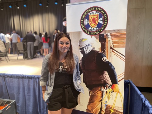 A womena stands next to a LIUNA stand up poster with a construction worker featured on it. She is standing at a job fair.