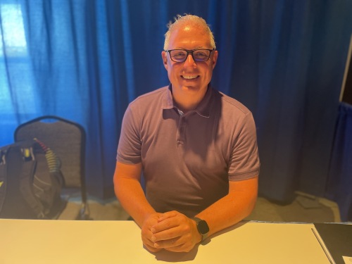 A man sits at a booth at a job fair with his hands in front of him, smiling. Behind him, there is a royal blue curtain.