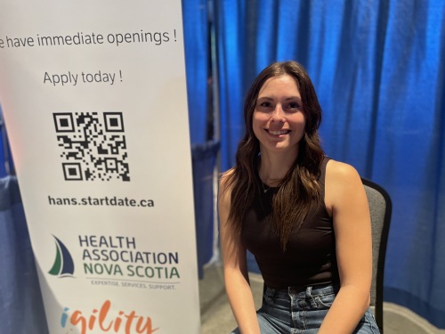 A woman sits in a chair next to a white stand up banner for Health Association Nova Scotia. There is a royal blue curtain behind her.