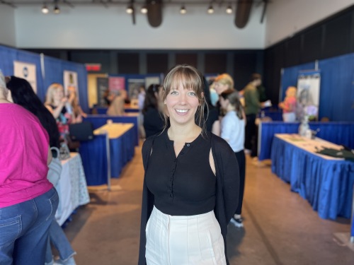 A woman stands in the middle of a job fair smiling. Behind here, there are tables draped in royal blue tablecloths and royal blue curtains.