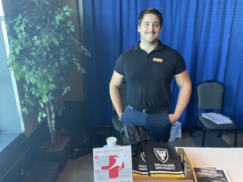 A man stands at a booth smiling at a job fair. Next to him is a potted tree and behind him is a royal blue curtain.