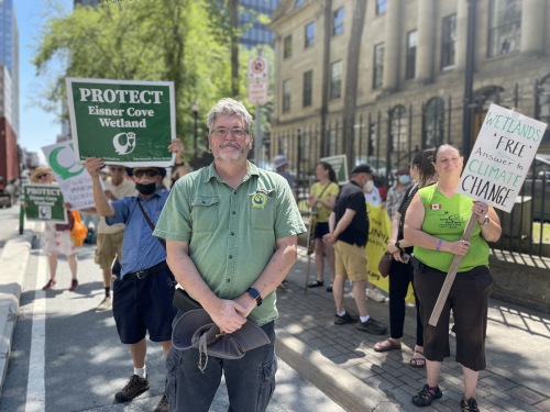 A man standing with enviromentalists protesting in the background with signs saying "protect the Eisner Cove Wetland."