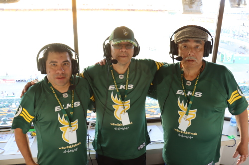 Three Indigenous men stand together wearing green and yellow Edmonton Elks jerseys and headsets in a broadcast studio. There is a stadium behind them.