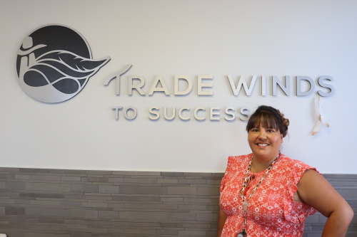 A woman standing in front of the Trade Winds sign black logo and white writing for "Trade Winds To Success."