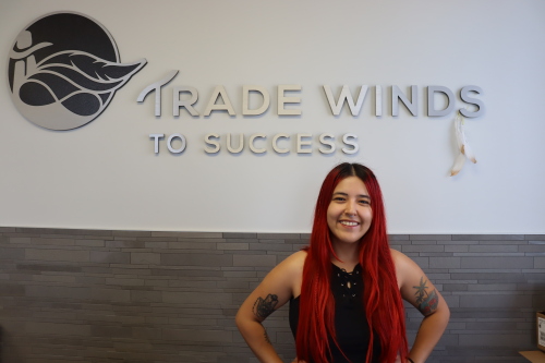 A female student with long red hair stands in front of the black and white Trade Winds To Success logo. 