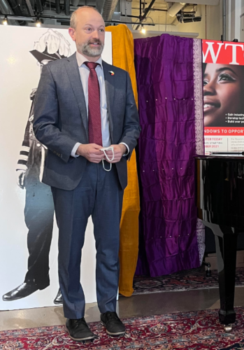A person in a suit with a red tie stands in front of a white poster with a drawn figure. Orange and purple drapes are on the left side behind.