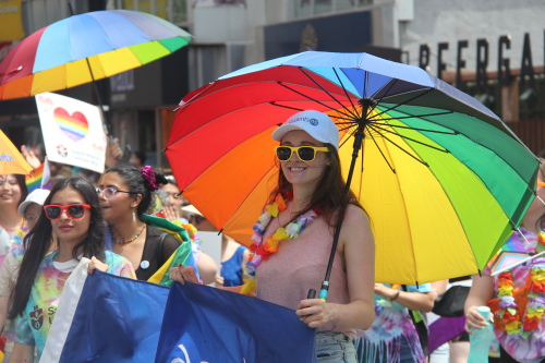 Photo of a woman at the pride parade holding an LGBTQ+ coloured umbrella, smiling at the camera.