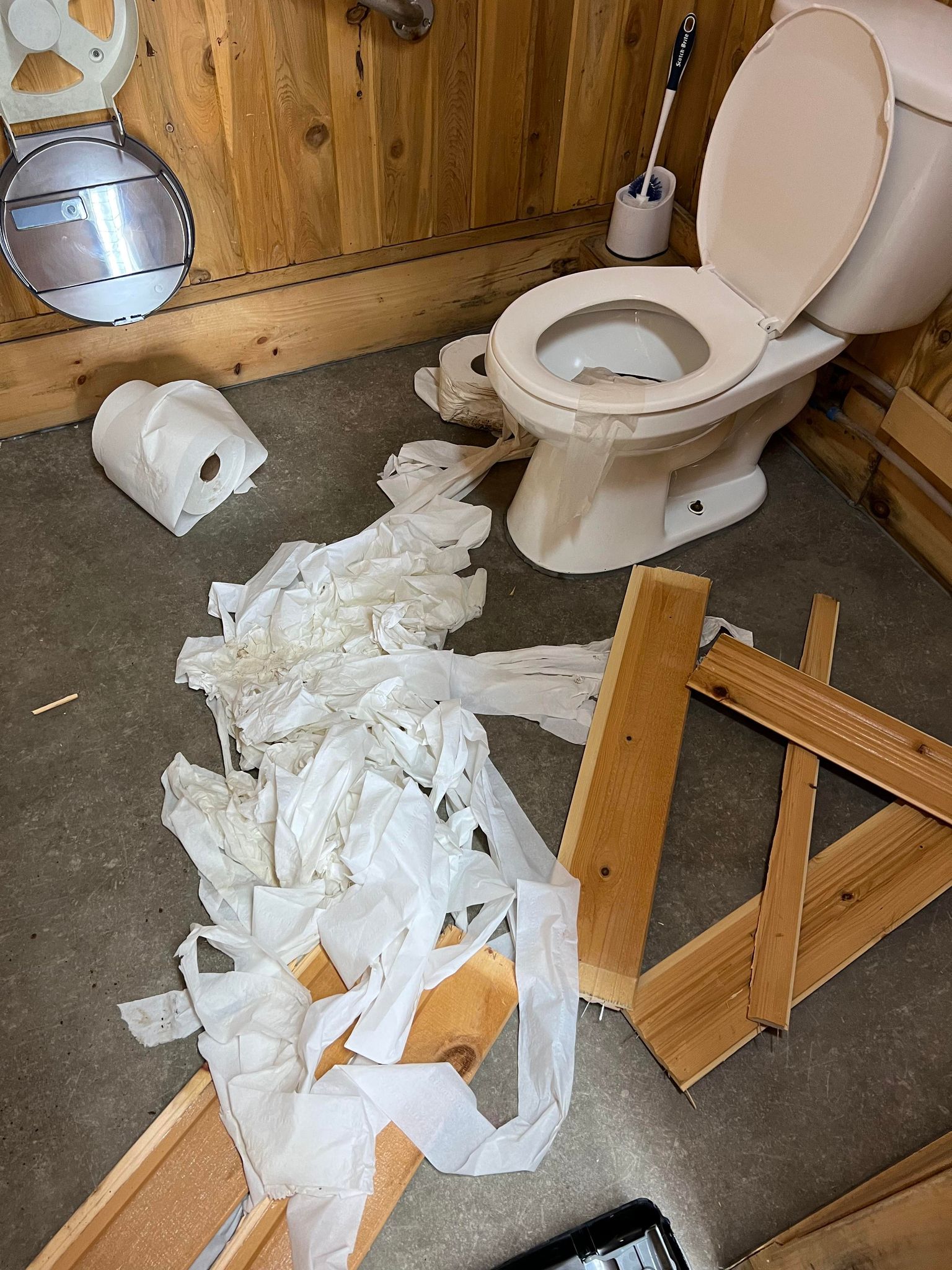 Papier de toilette étendu sur le sol d'une salle de bain publique. Plusieurs morceaux de bois cassé traîne au sol.