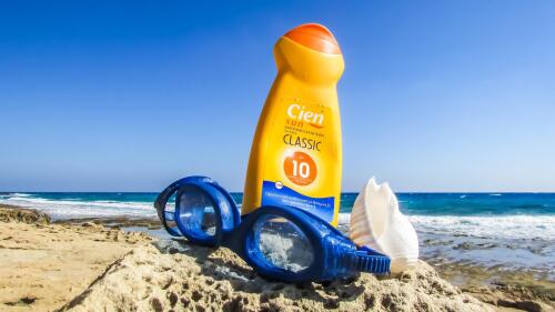 Sur une plage, une paire de lunettes de baignade bleue posée devant un flacon de crème solaire.