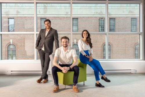 A professional photo of three people. One man is standing and leaning on a chair while another man sits in the chair and a woman sits on the back of the chair. There is a panel of windows and a brick building behind them.