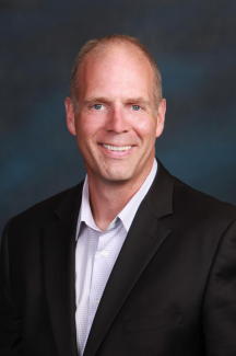 A headshot of a smiling man. He is wearing a black suit and white collared t-shirt. There is a dark blue background behind him.