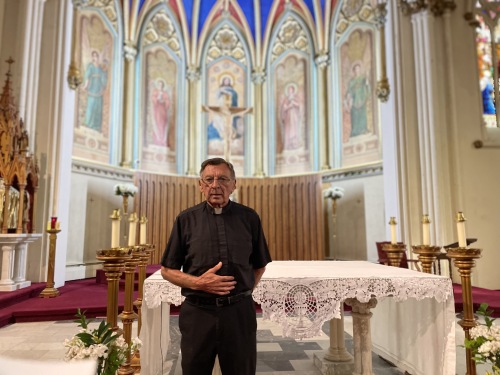 A priest stands near the restored murals inside the church. He stands in front of an alter.