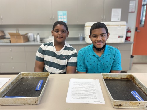 Photo of two young brothers sitting at a table smiling. There is a scalpel and tray on the lab table.