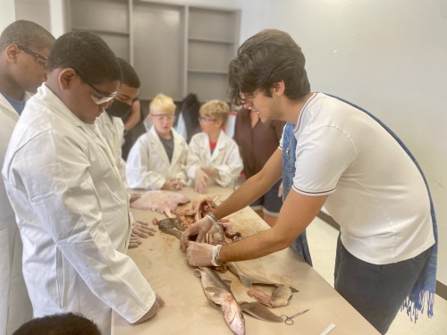 Photo of a man holding a shark on the table. There are kids in labcoats and goggles watching in the background.