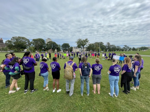 About 30 people dressed in purple shirts that say International Overdose Awareness Day gathered in a circle in the Halifax Commons park. They are standing on grass. There are cloudy skies above them. Many of them are holding up different coloured signs.