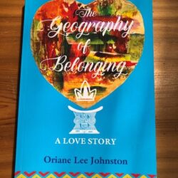 A multi-coloured book cover sits atop a wooden table top. It reads "The geography of belonging: a love story. Oriane Lee Johnston."