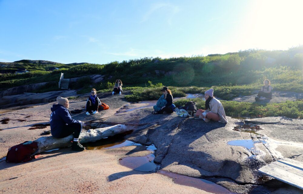 Un groupe de personnes assises sur un plaqué rocher écoute une femme qui lit.