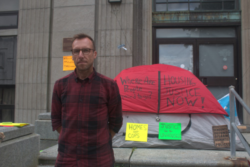A male volunteer stands in front of a grey and red tent at a rally. The tent has multicoloured signs on it.