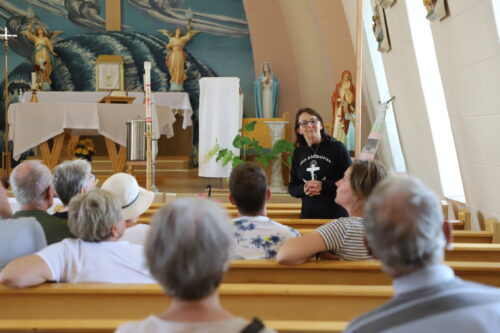 Dans des bancs d'église, des gens, de dos, écoutent une dame portant un chandail de Tête-à-la-Baleine parler.