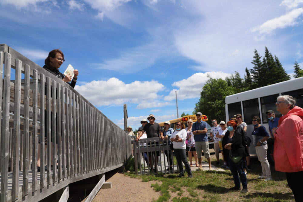 Sur une galerie en hauteur, une dame montre un livre à une foule attentive. Derrière, des minibus sont stationnés.