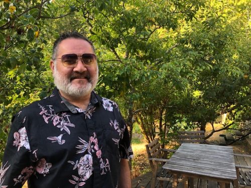 A man in sunglasses sporting a beard and wearing a floral patterned shirt stands in front of a treed background.