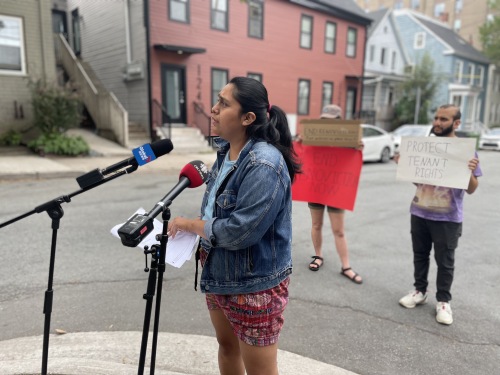 Woman in a jeans jacket with her hair in a pony tail is speaking in to two microphones, there are two people in the back holding posters to support tenant rights.