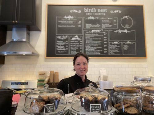 A woman is smiling inside a cafe. There is a menu on a chalkboard with Bird's Nest food and drinks. She is standing behind a platter of cookies and muffins as she smiles for the photo.