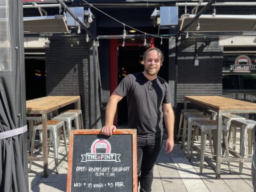 Photo of a man standing outside The Pint Public House, there are tables on the background and a chalk board with the Pint's specials. He is smiling.