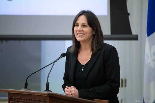 Pictured is Isabelle Charest standing behind a podium speaking at a conference in Brome-Missisquoi. She is wearing a black t-shirt with a black blazer.