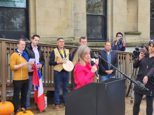 La chef du parti libéral du Nouveau-Brunswick a pris la parole devant l'assemblée législative. Elle parle au pupitre entourée de membres de la SANB portant des drapeaux Acadiens.