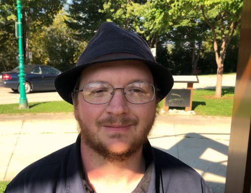A man in a hat and glasses stands outside.