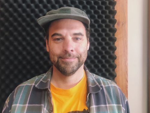 A man in a hat and plaid shirt smiles for a photo.