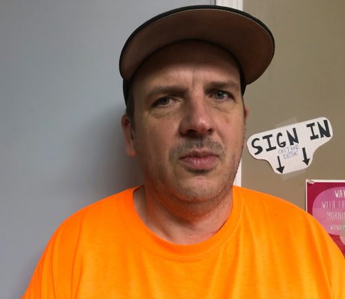 A man in an orange shirt and hat stands against a wall.