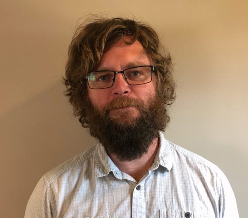 A bearded man with tousled brown hair wears a white collared shirt.