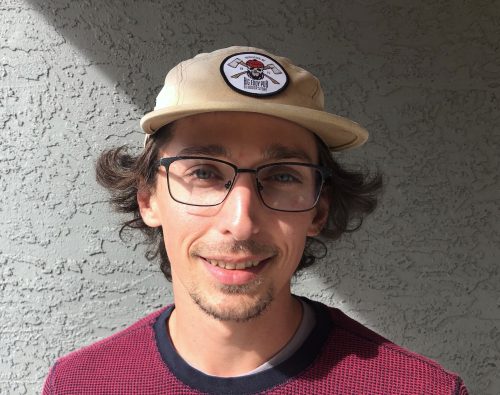 A man with brown hair and glasses wearing a hat stands against a wall.