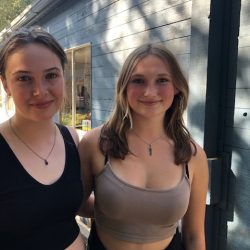 Two teenage girls wearing black and beige tops stand beside a blue coloured building.