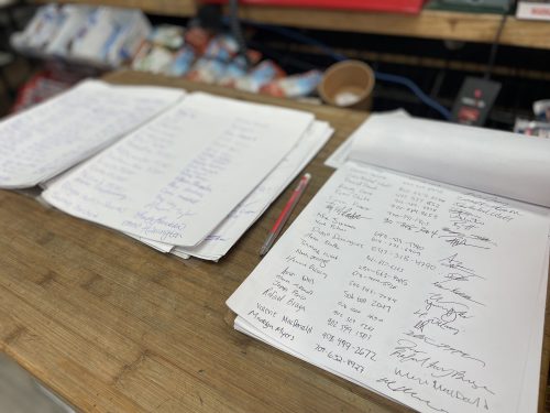 Photo of signatures on papers. There are three rows of papers filled with signatures. They are on a table in a convenience store, there are snacks in the background.