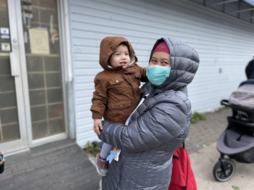A woman in a coat holding her baby daughter in her hand. She is wearing a mask, and her kid is smiling at the camera.